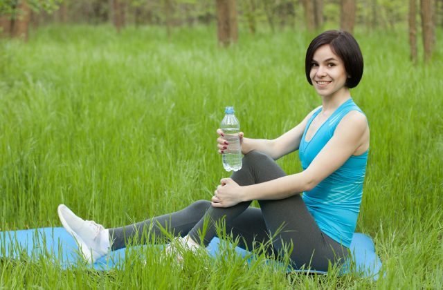 Woman in blue tank top sitting on grass with a yoga mat, holding a water bottle, enjoying outdoor exercise