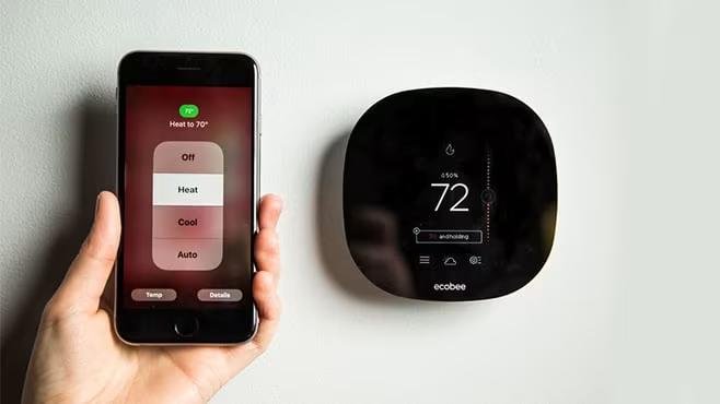 A person holds a smartphone beside a smart thermostat, demonstrating smart home technology integration