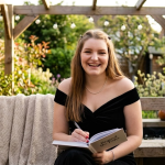 Woman in black dress smiling, writing in notebook on garden bench under pergola with plants surrounding her