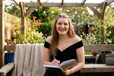 Woman in black dress smiling, writing in notebook on garden bench under pergola with plants surrounding her