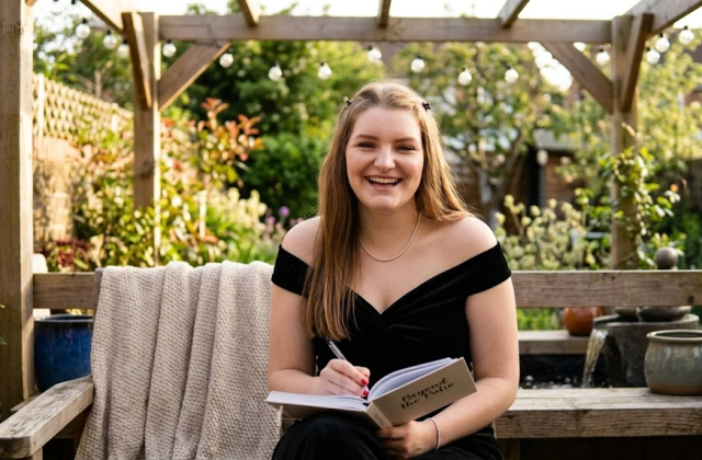 Woman in black dress smiling, writing in notebook on garden bench under pergola with plants surrounding her